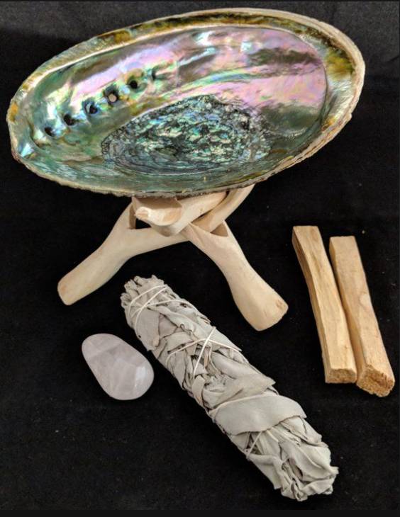 An abalone shell, white crystal, White Sage Smudging kit, and pieces of palo santo wood arranged on a black background.