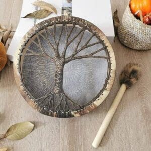 Decorative round Shaman Drum “Tree of Life” resting on a wooden surface alongside autumn leaves and a drumstick.